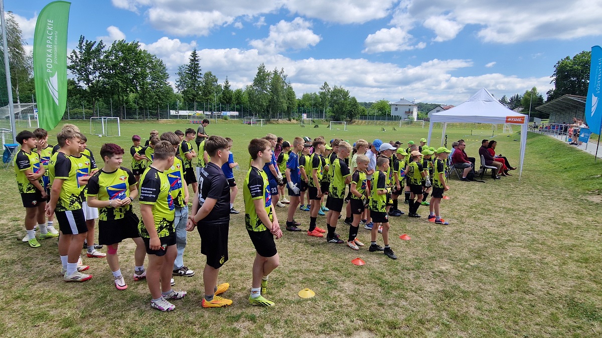 Grupa młodych piłkarzy w jaskrawych strojach czeka na boisku sportowym. W tle widoczni kibice i drzewa.